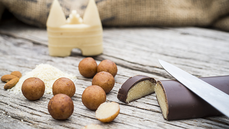 Ein Arrangement von Lübecker Marzipan auf einem rustikalen Holztisch. Im Vordergrund liegt ein Marzipanbrot, halb aufgeschnitten, neben einem Messer. Rundherum sind Marzipankugeln und Mandeln verstreut und im Hintergrund steht eine kleine Marzipanfigur in Form des Holstentors.